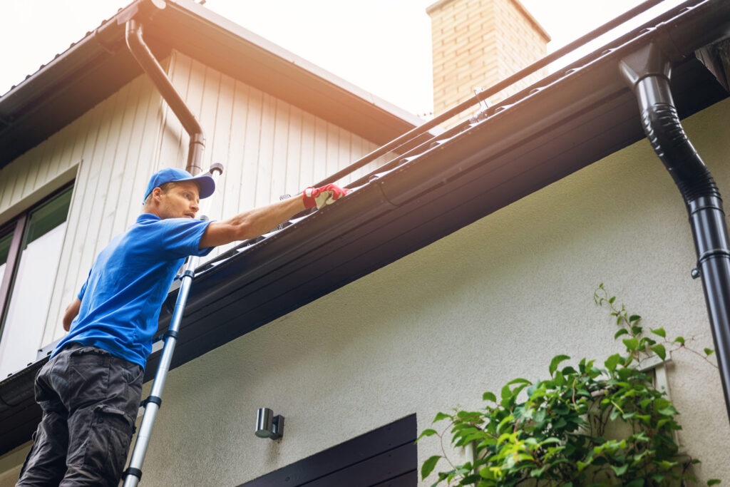 A man on a ladder leaning against the roof of a house, cleaning the gutters